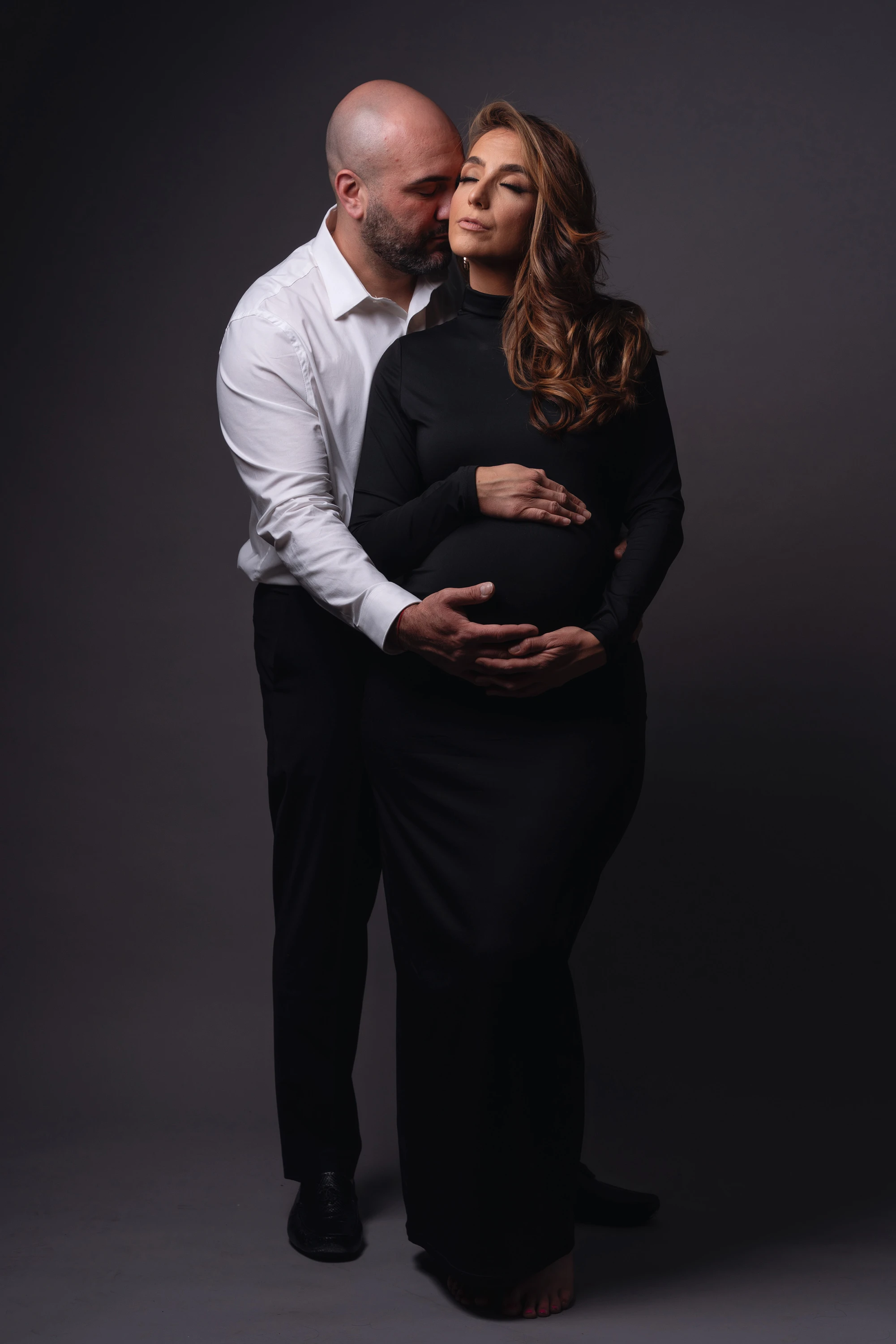 man in business casual attire hugging his pregnant wife in front of dark backdrop