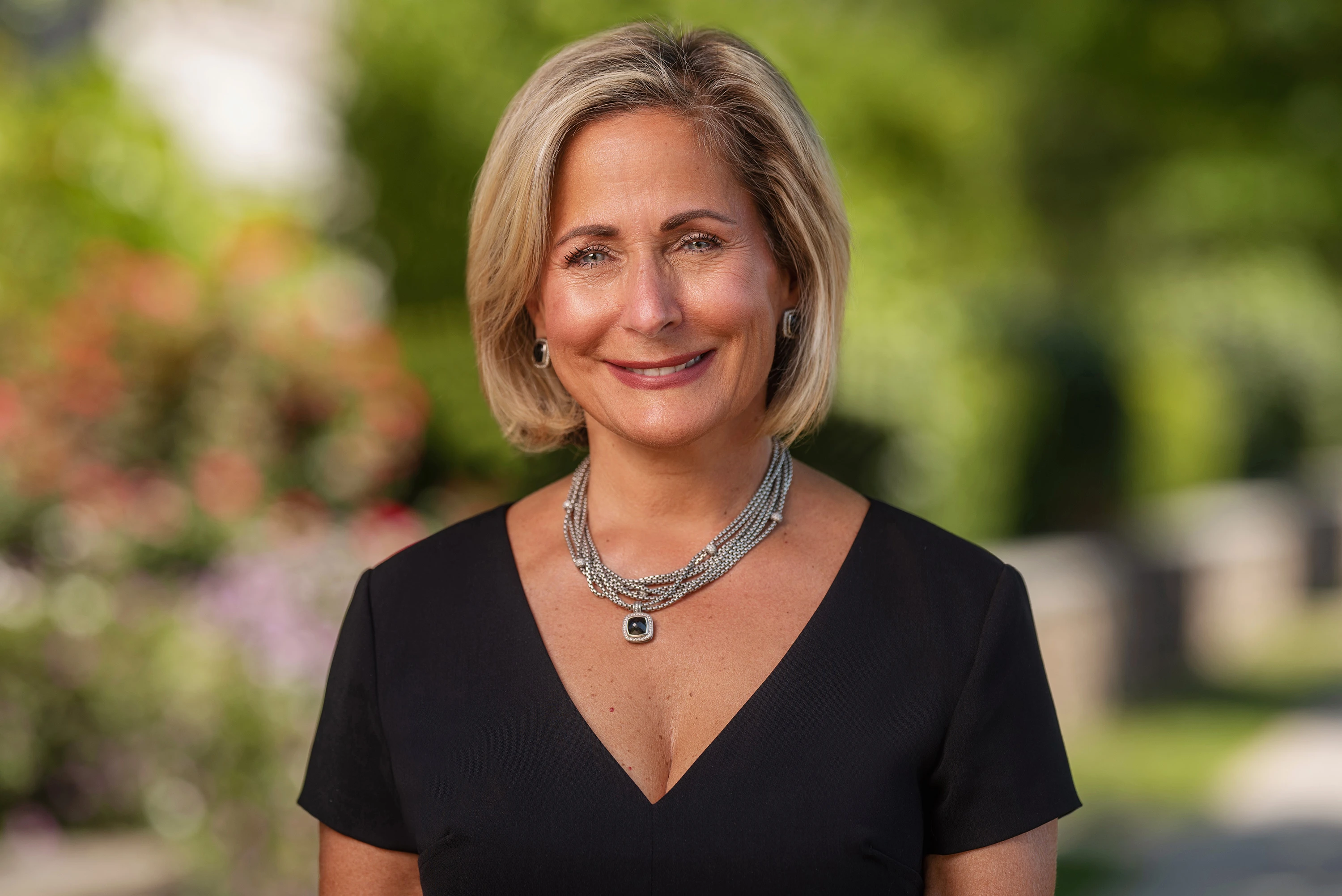 close up of blonde haired woman with necklace and black dress shirt posing outdoors with blurred background for headshot